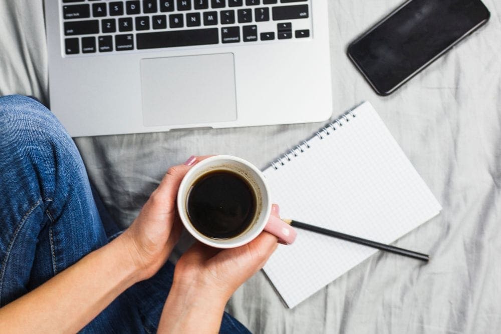 Tasse de café tenue en main avec un carnet et un crayon posés près d’un ordinateur portable – image de la newsletter Kaellyana.