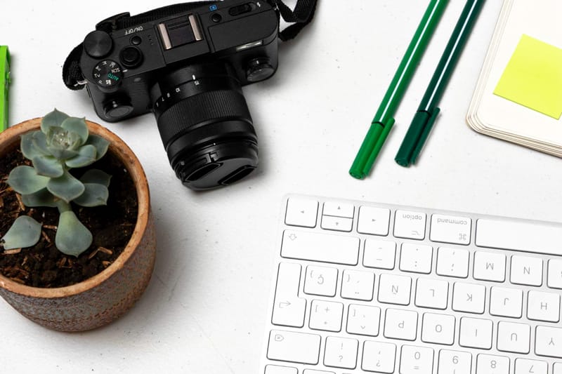 Appareil photo noir avec un clavier, des stylos verts et un carnet sur un bureau blanc – Illustration d’un article sur les banques d’images thématiques.